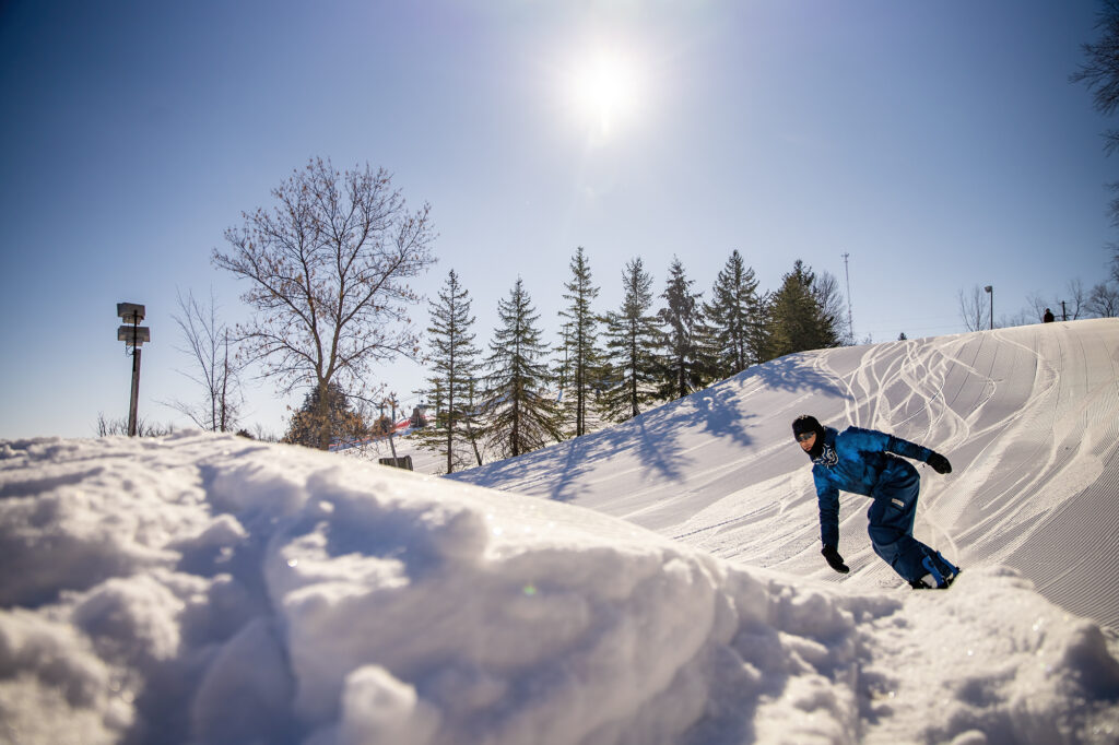 A snowboarder approaches a snowy jump on a ski slope.